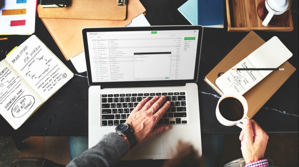 Seorang pria sambil memegang cangkir kopi tengah membuka email di laptop